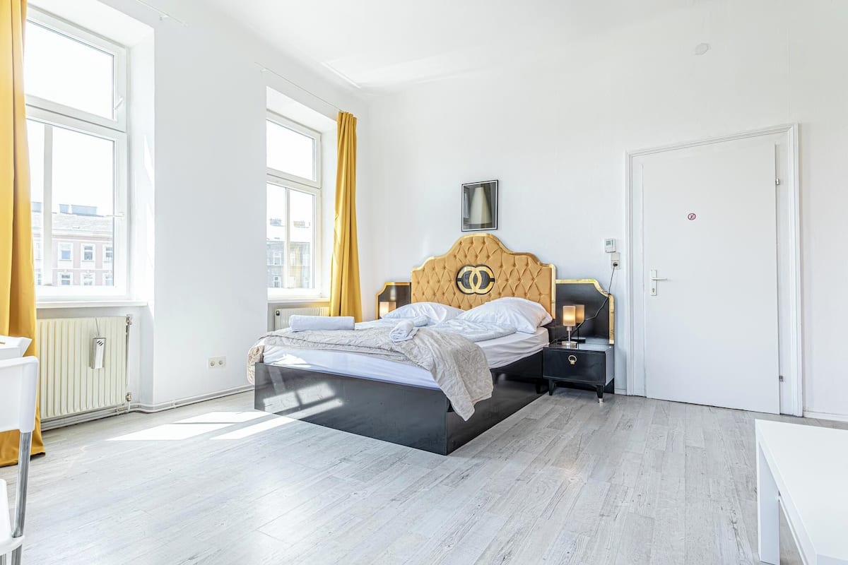 A modern bedroom with a large bed, a nightstand, a bedside table, a radiator, and a window with curtains.