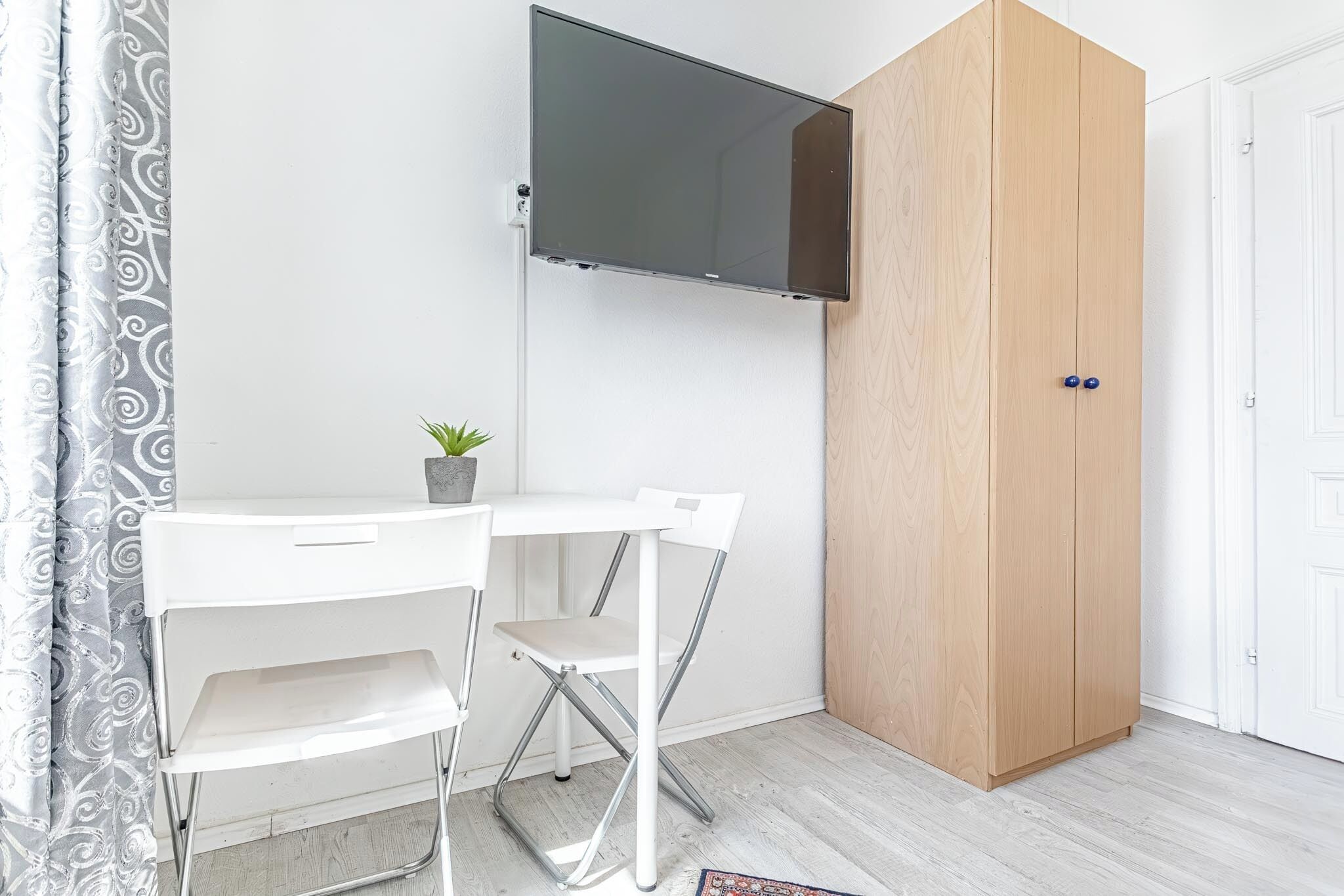 A compact room with a white desk, a foldable chair, a TV mounted on the wall, and a wooden wardrobe.