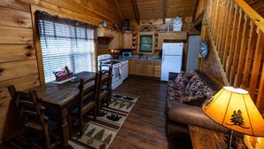 Interior acogedor de una cabaña con una mesa de madera, sillas, un sofá y una cocina con estufa, microondas y refrigerador.