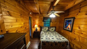 Un dormitorio de cabaña de madera con cama, mesita de noche, lámpara, ventilador de techo y un cuadro en la pared.