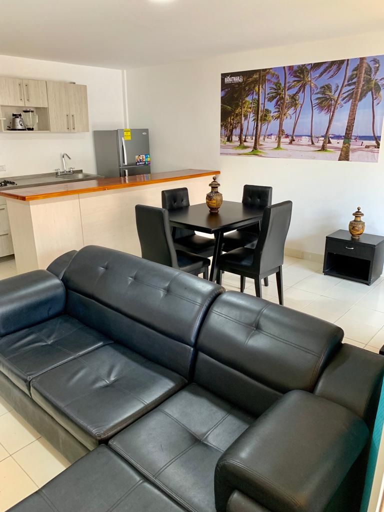 A modern living room with a black leather sofa, a dining table with chairs, a kitchen area, and a beach-themed wall art.