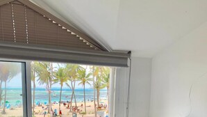 Ein Schlafzimmer mit Bett, Nachttisch, Lampe und Fenster mit Blick auf einen Strand und Palmen.