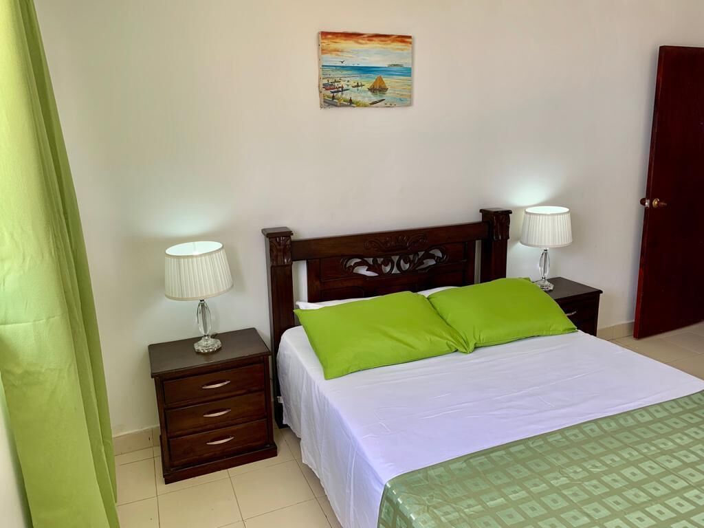 A bedroom with a wooden bed, green pillows, a white bedspread, two bedside tables with lamps, a nightstand, and a painting on the wall.
