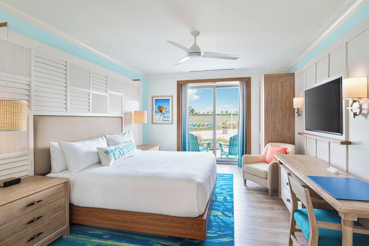 Una habitación de hotel con una cama grande, un escritorio con televisor, un balcón con sillas y vista a la playa.