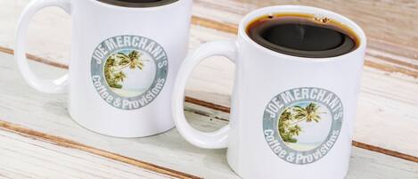 Deux tasses à café blanches ornées d’un motif tropical et d’une inscription, posées sur une surface en bois.