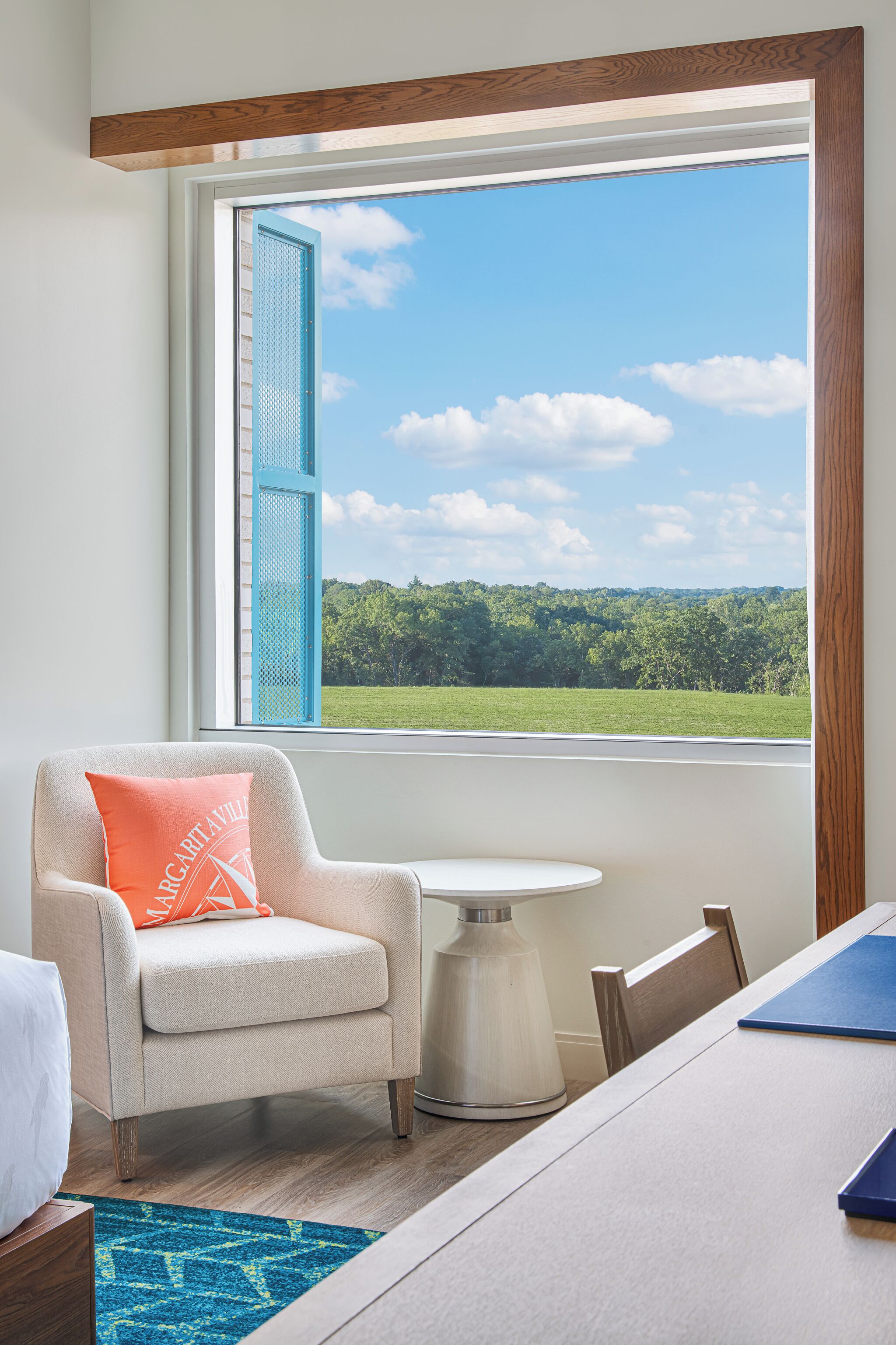 A room with a view of a green field through a window with blue shutters. There is a white armchair with an orange pillow, a small white table, and a wooden chair.