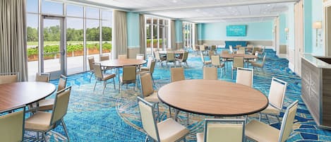 A hotel conference room with round tables, chairs, and a large window offering a view of greenery.