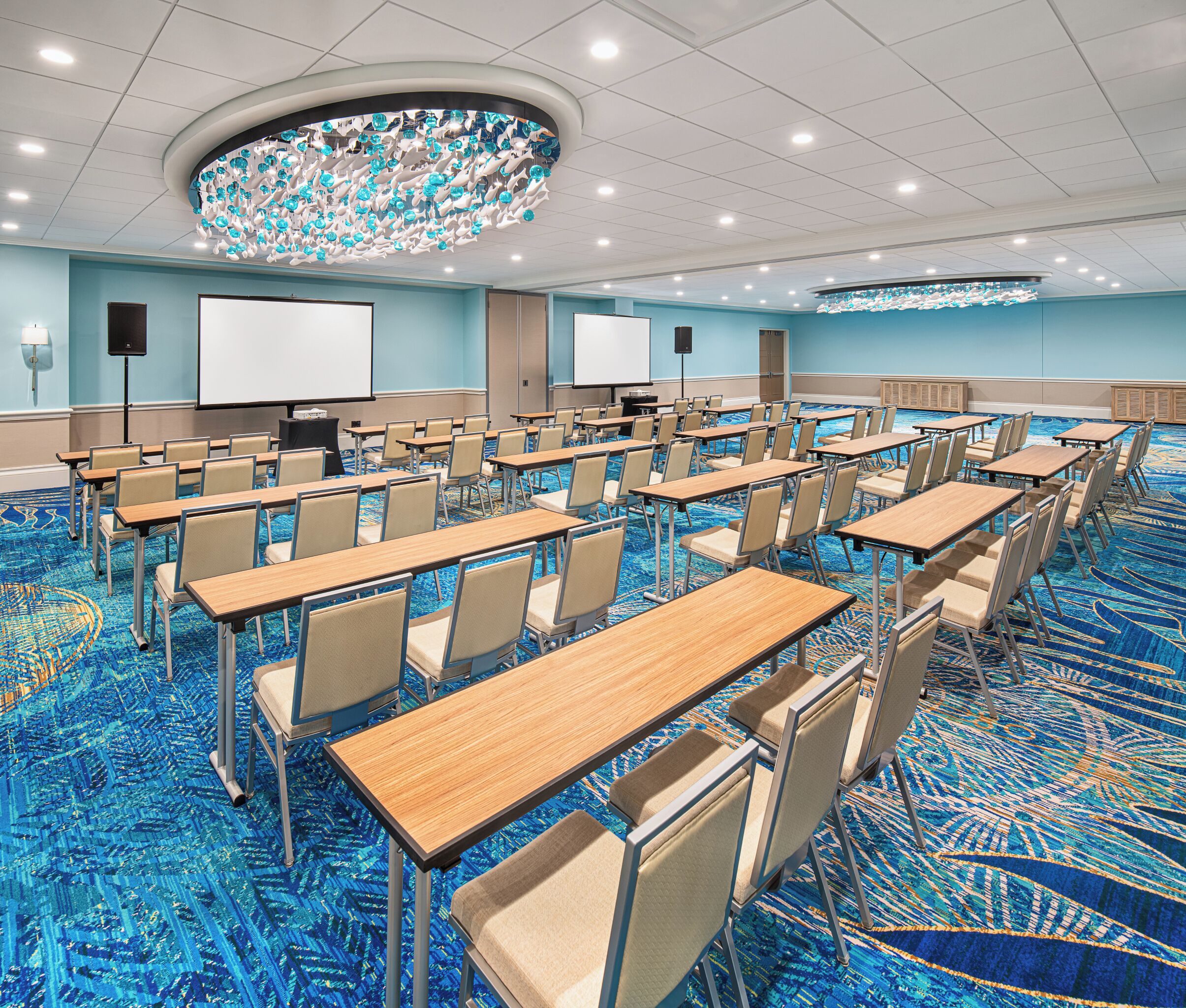 A conference room with rows of tables and chairs, a large screen, and a ceiling light fixture.