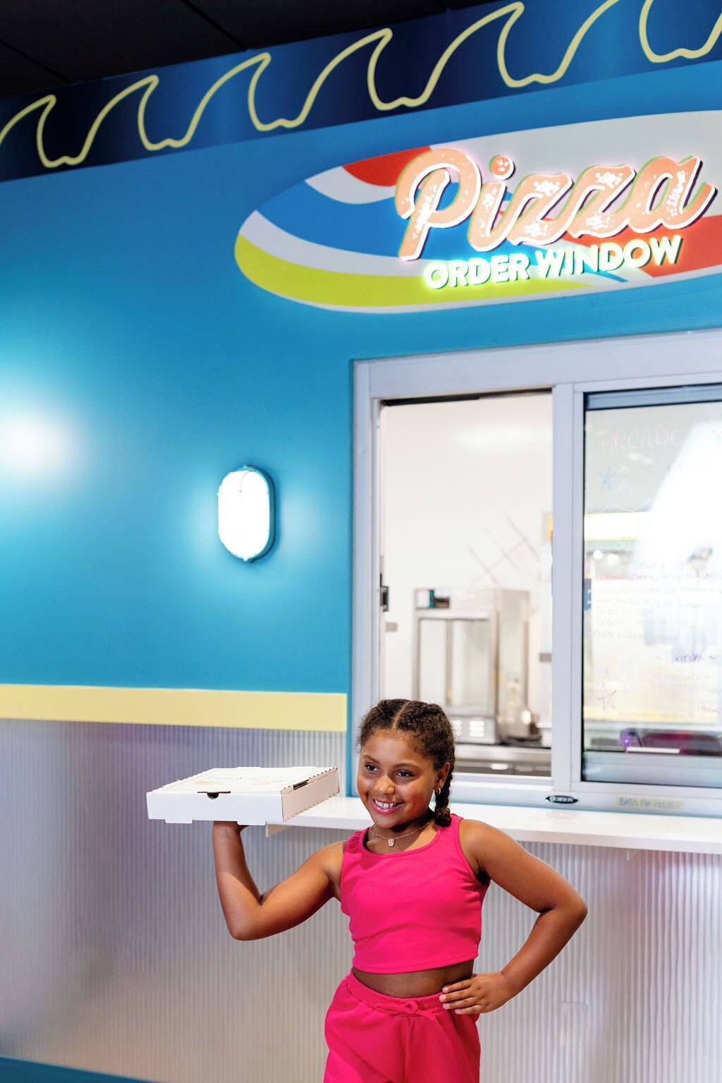 A person holding a pizza box in front of a brightly colored wall with a 'Pizza Order Window' sign.