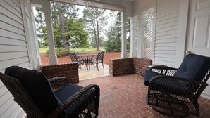 Une véranda avec des fauteuils en osier et une table, un sol en briques et une vue sur une pelouse et des arbres.