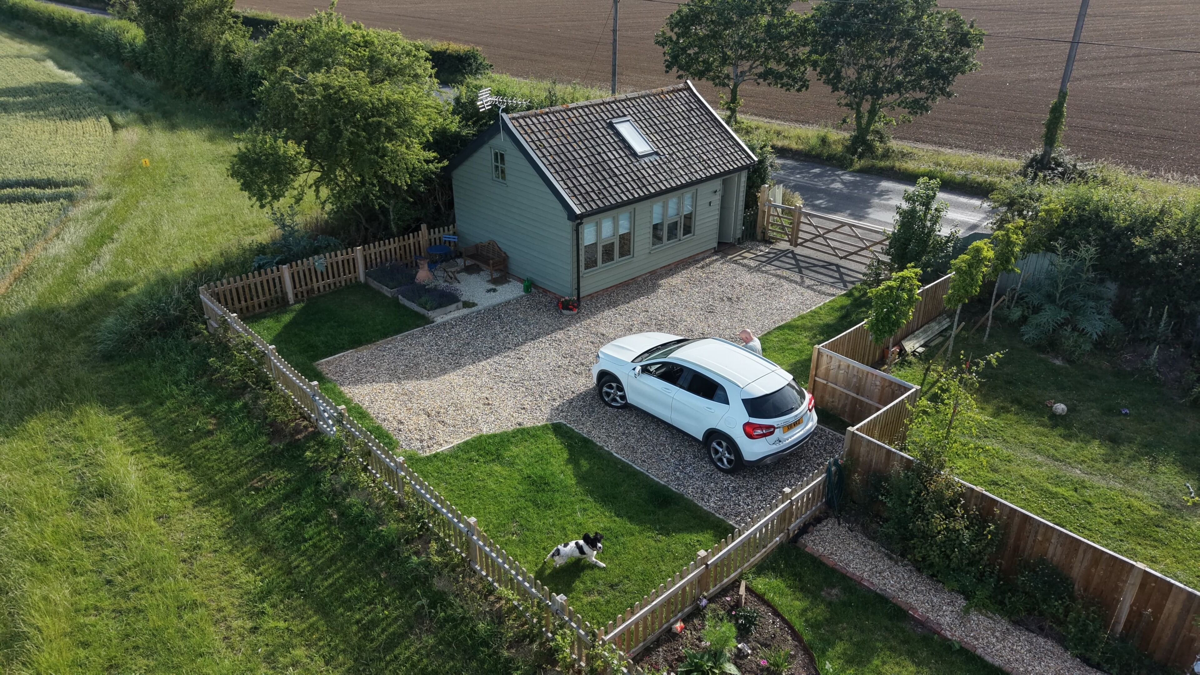 Une maison avec une voiture garée dans l’allée, entourée d’une cour clôturée et d’un chemin de gravier.
