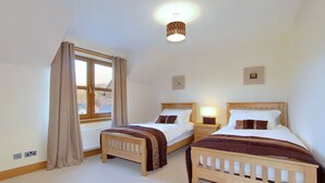 A bedroom with two wooden beds, a window with curtains, a ceiling light, and a small bedside table with a lamp.