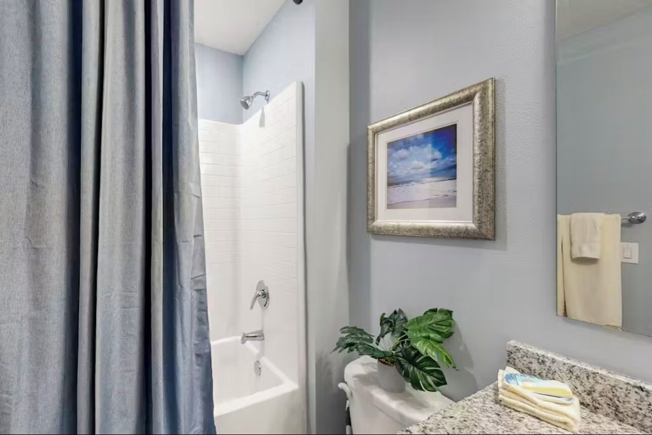 Une salle de bain avec une douche, un lavabo avec un miroir, un tableau encadré au mur et une plante posée sur le comptoir.