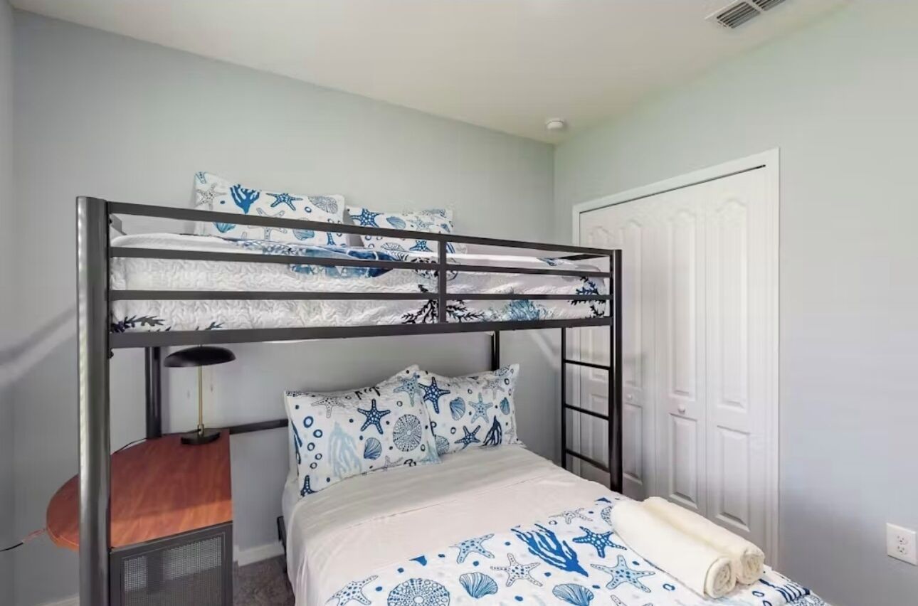A bunk bed with a white and blue nautical-themed bedding, a bedside table with a lamp, and a white door.