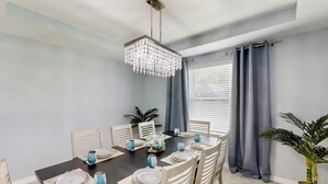 A dining area with a dark wooden table set for six, white chairs, a chandelier, and a potted plant.