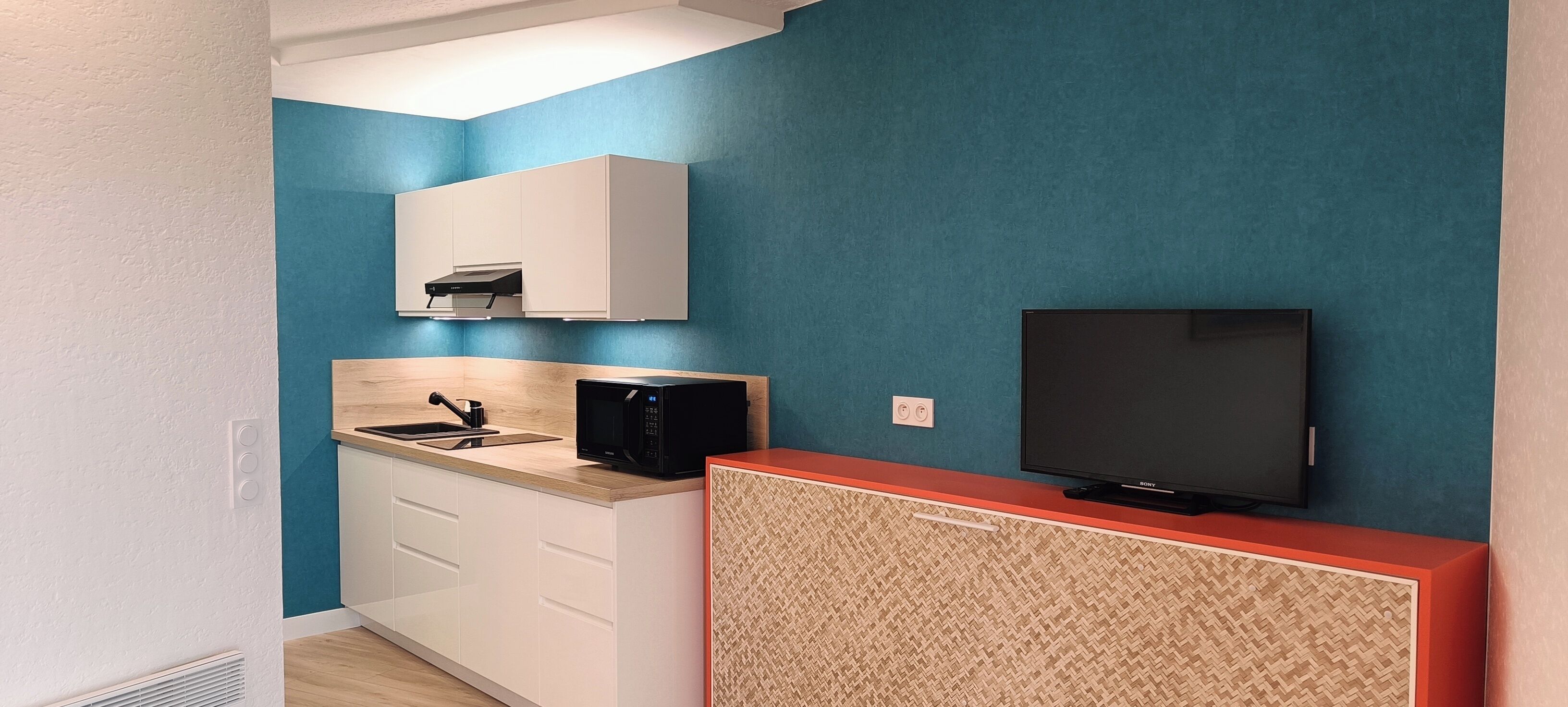 A compact kitchen with a microwave, sink, and a flat-screen TV on a stand.