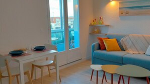 Un salon avec un canapé bleu, une table à manger avec des chaises et une fenêtre donnant sur des immeubles.