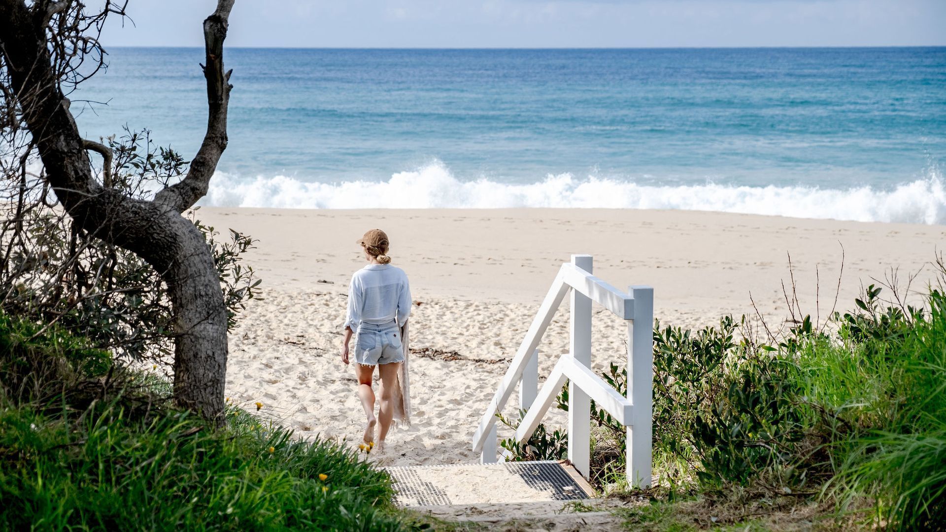 A beach with a person walking towards the sea, a white wooden structure, and a tree.
