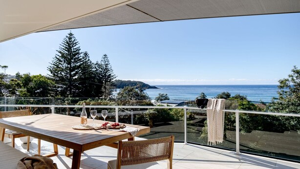 Ein Holztisch mit Flasche und Gläsern, eine gewebte Bank und Blick auf das Meer und die Hügel.