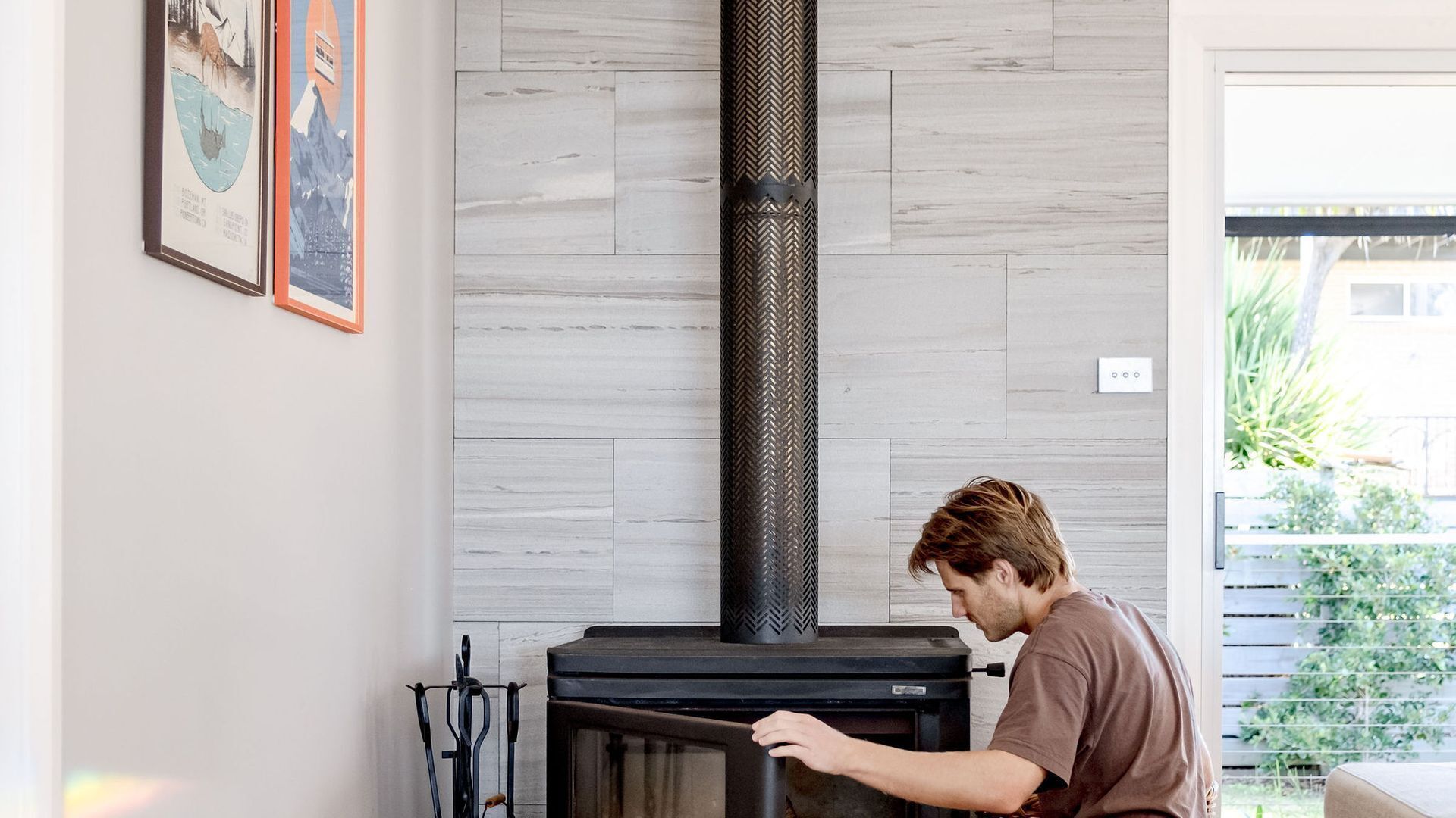 A person operating a wood stove in a room with a herringbone-patterned chimney, light wooden walls, and a framed picture on the wall.
