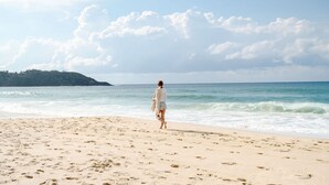 Ein Strand mit Fußabdrücken, die zum Wasser führen, eine Person, die vom Betrachter wegggeht, und eine entfernte Baumgrenze.