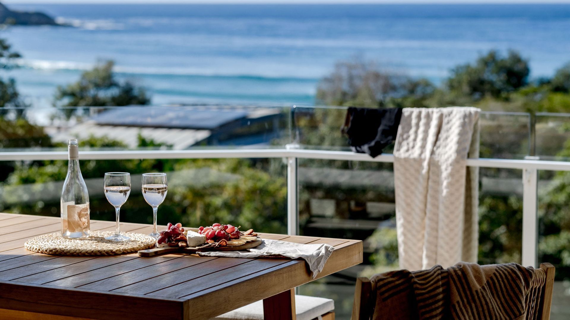A wooden table with a bottle, glasses, grapes, and a cheese board, overlooking a coastal view with a balcony and a hanging towel.
