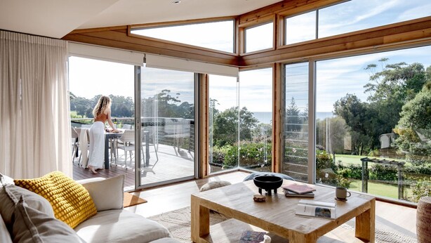 A spacious living room with a large sliding glass door leading to an outdoor deck, a wooden coffee table, and a sofa with a yellow cushion. - Everlong Mollymook - Spacious Home on Beachside Golf Course with Ocean views (Mollymook)