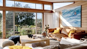 A spacious living room with a large window, a brown leather sofa, and a painting of a sailboat. - Everlong Mollymook - Spacious Home on Beachside Golf Course with Ocean views (Mollymook)