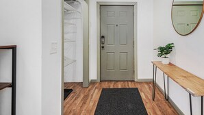 A hallway with a wooden floor, a grey door, a small table with a plant, and a mirror on the wall.