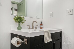 Ein Badezimmer mit weißem Waschbecken, schwarzem Waschtisch, einer Zimmerpflanze, einem Handtuch und einem Toilettenpapierhalter.