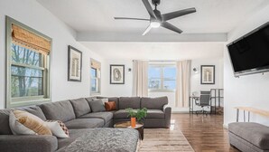 A spacious living room with a grey sectional sofa, a coffee table, and a TV mounted on the wall. There is a ceiling fan, a window with blinds, and a desk with a chair in the background.