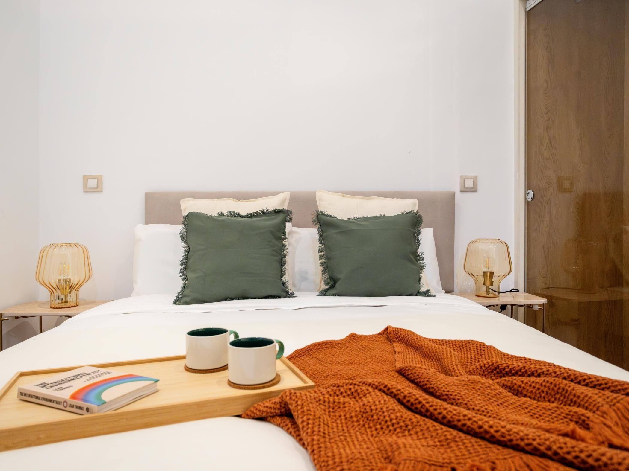 A neatly made bed with two pillows, a bedside table with a lamp, and a tray with books and mugs.