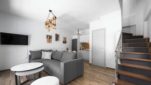 Una sala moderna con un sofá gris, una mesa de centro redonda y un reposapiés blanco. Una escalera de madera conduce a una zona de cocina.