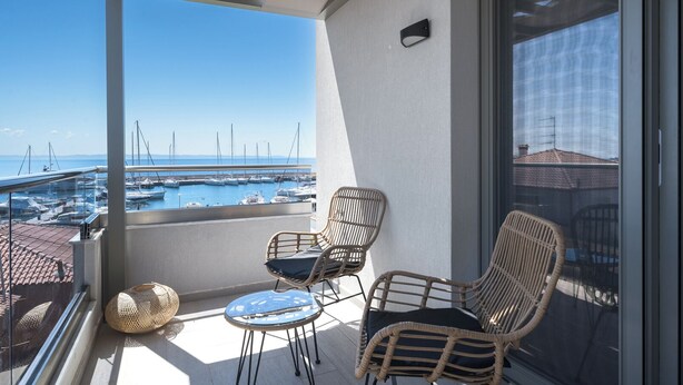 Un balcón con sillas de mimbre y una pequeña mesa, con vista a un puerto deportivo con barcos.