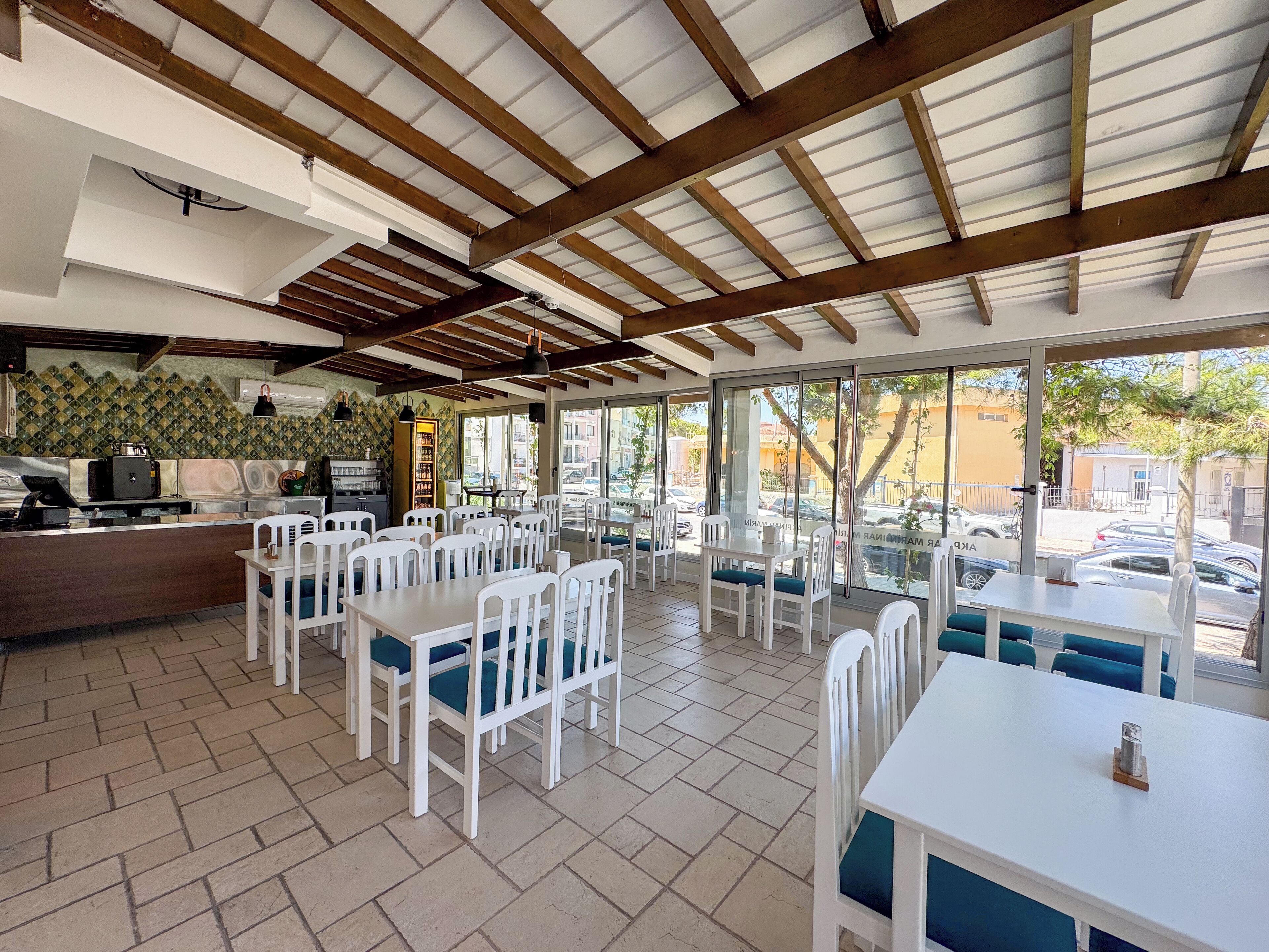 Un restaurante con piso de baldosas, mesas y sillas blancas, una barra con una máquina de café y vistas a la calle a través de amplias ventanas.