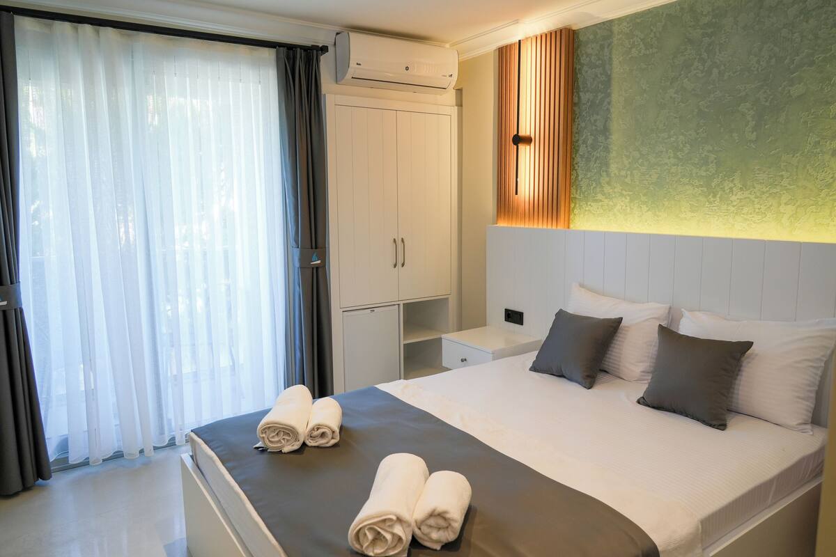 Een hotelkamer met een bed, nachtkastje, kledingkast en een groot raam met gordijnen.