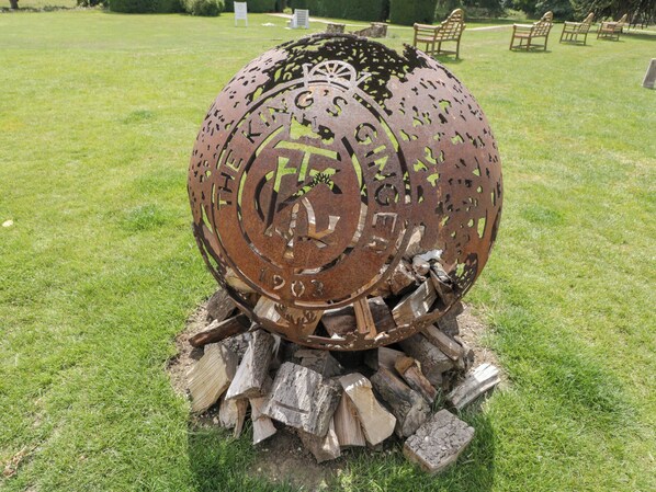 Une sculpture métallique représentant une roue et des chiffres, placée sur un champ d’herbe, avec des troncs d’arbres en dessous.