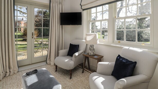 Ein Wohnzimmer mit einer Couch, einem Sessel und einem kleinen Tisch mit Lampe. Ein Fernseher ist an der Wand angebracht und es gibt ein Fenster mit Vorhängen.