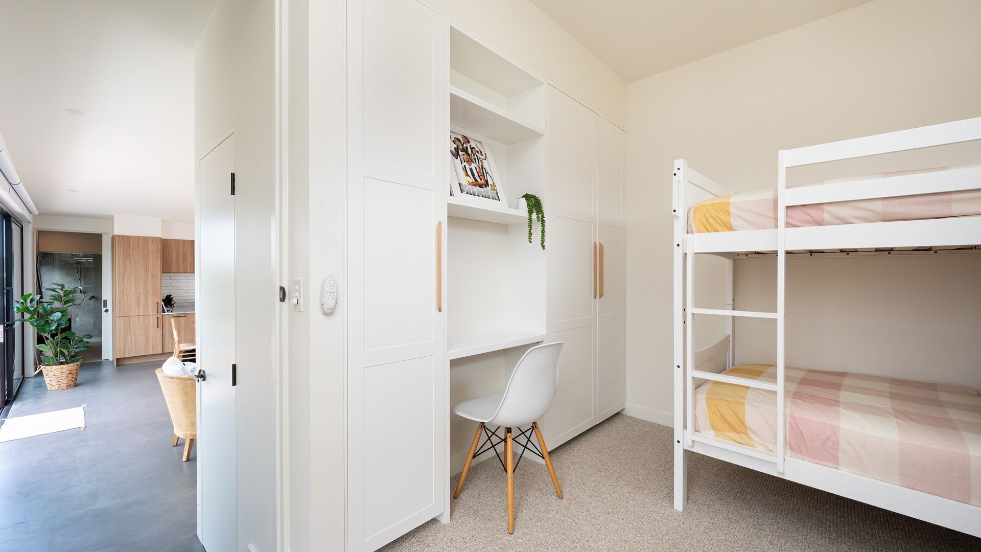 Une chambre moderne et bien éclairée, avec un lit superposé, un bureau blanc et une chaise. On y trouve également un pot de plante et un panier posés sur le sol.