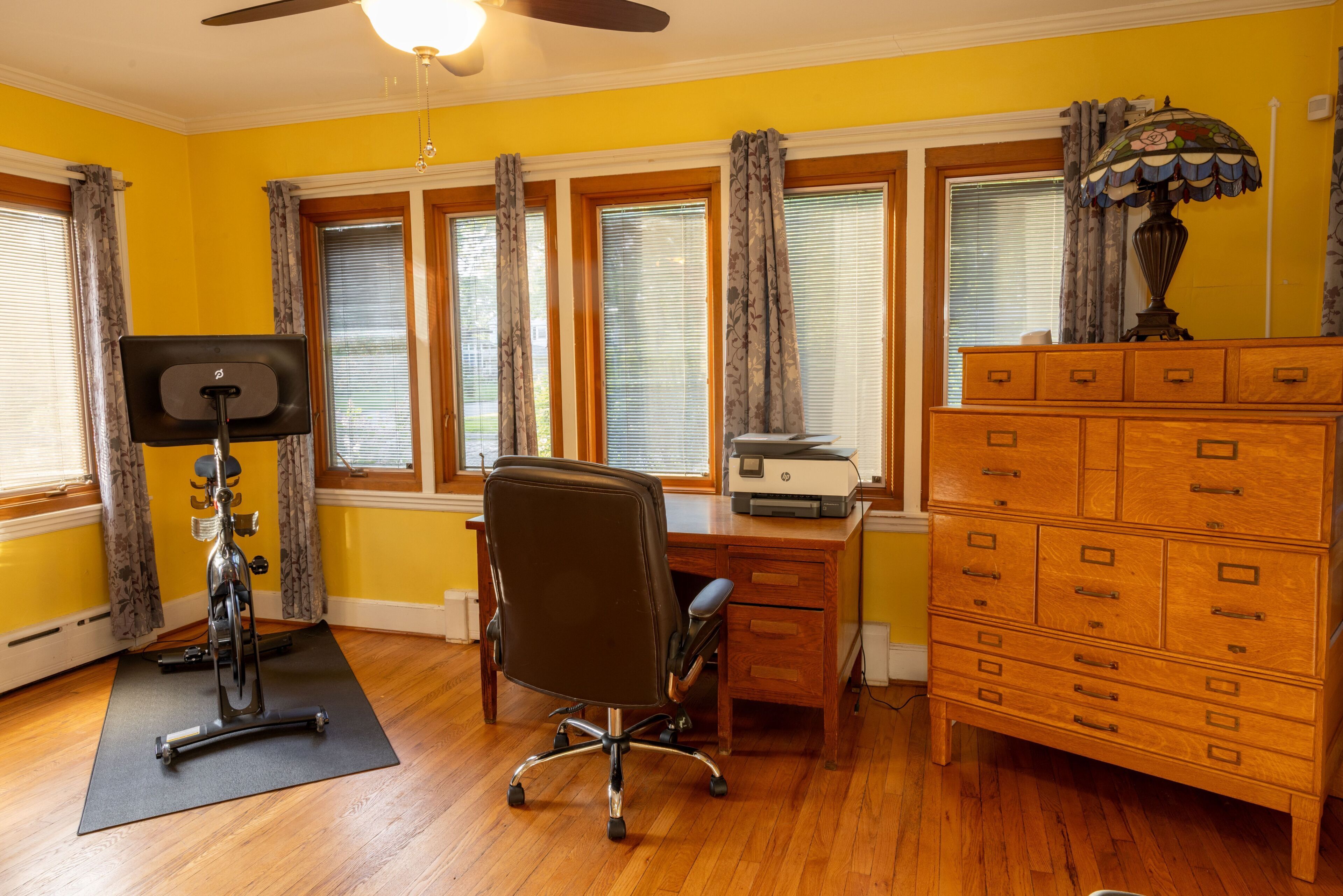 A room with a wooden desk, an HP printer, a chair, a treadmill, and a dresser.