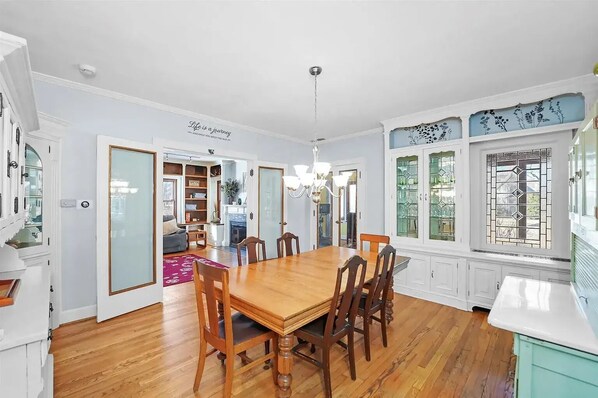 Une salle à manger avec une table et des chaises en bois, un lustre et une vue sur un jardin, visible à travers une fenêtre ornée de vitraux décoratifs.