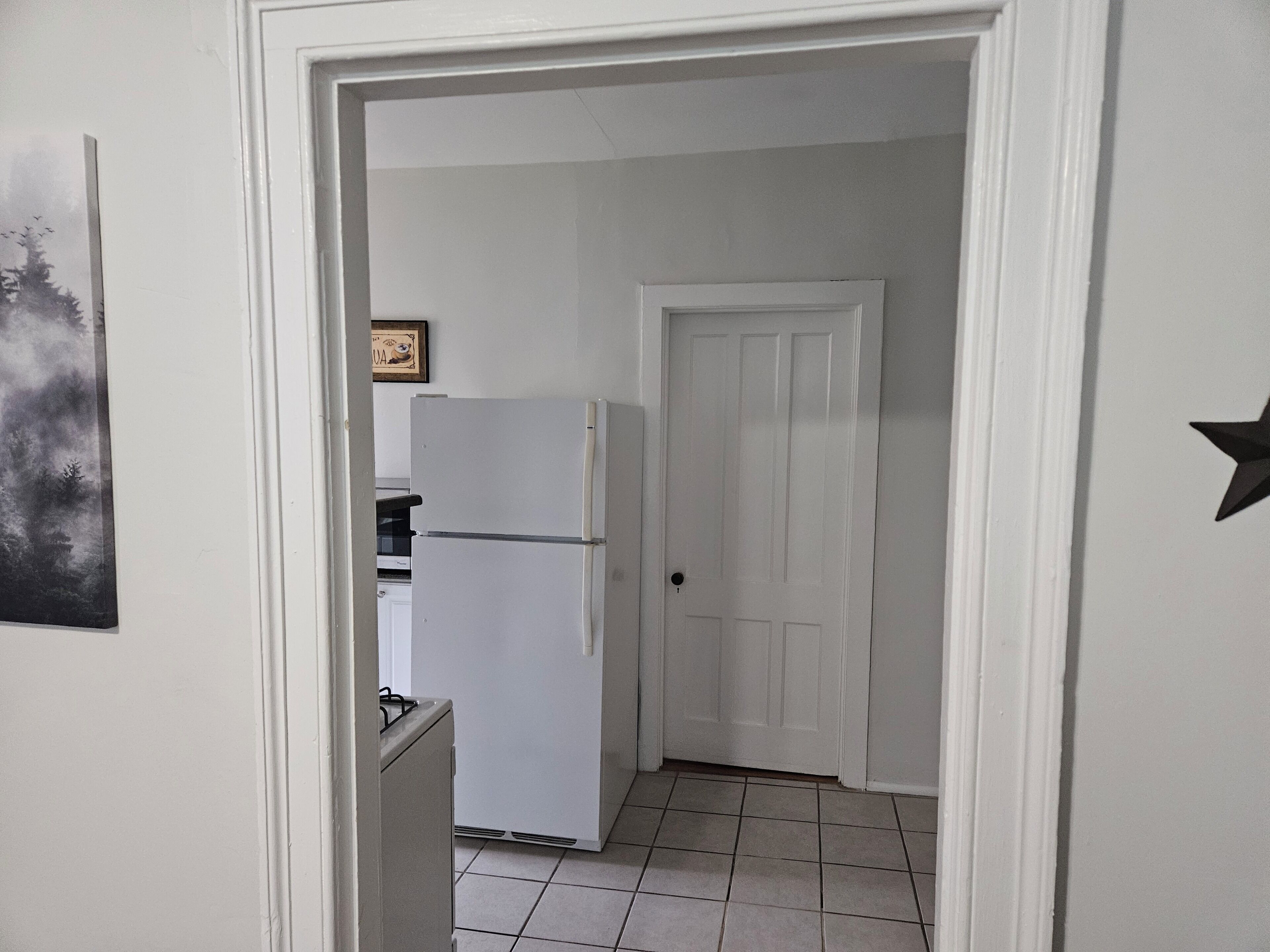 A narrow hallway with a white door at the end, a white refrigerator, and a tiled floor.