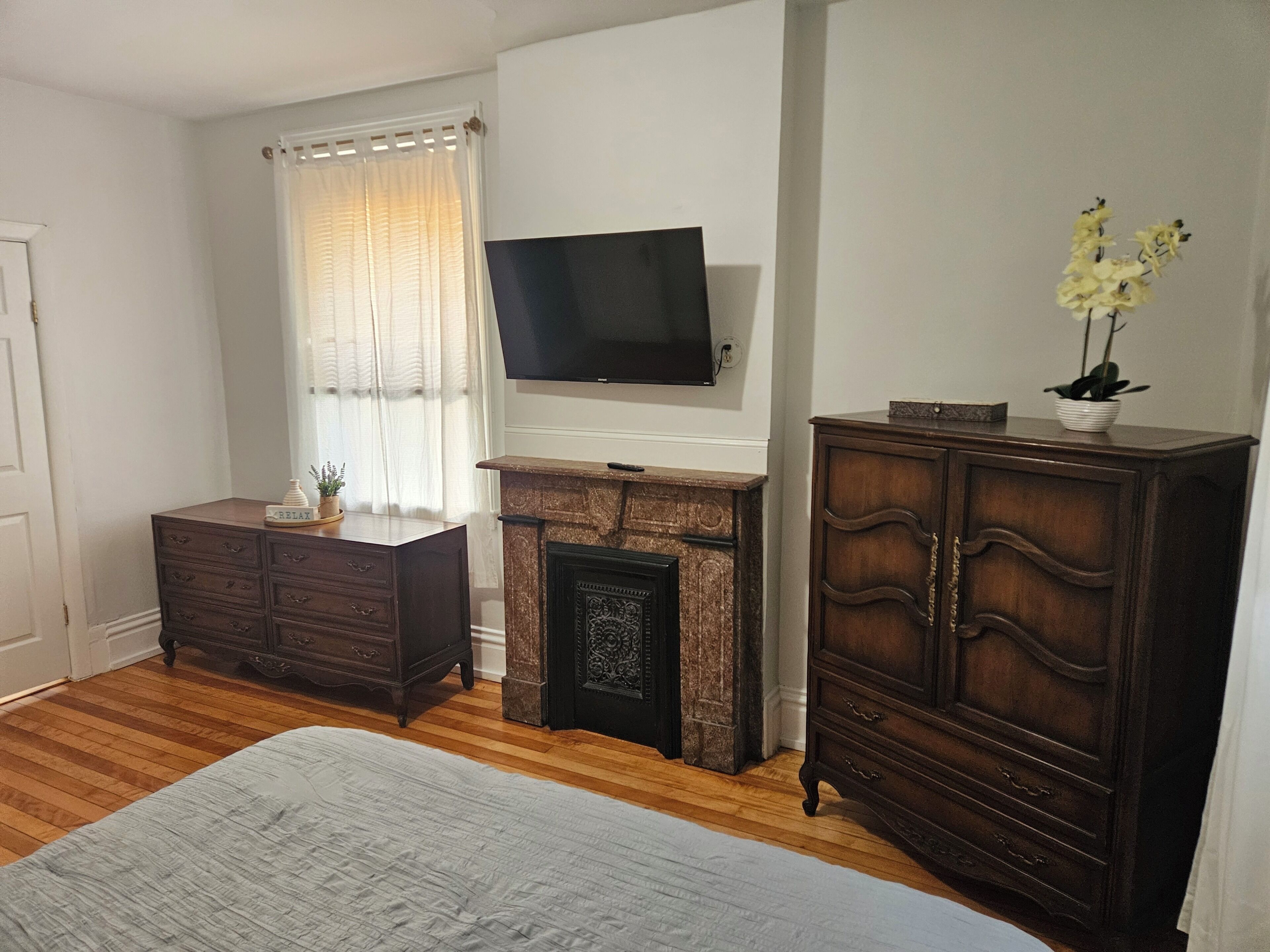 A bedroom with a bed, a dresser, a television, a fireplace, and a window with curtains.