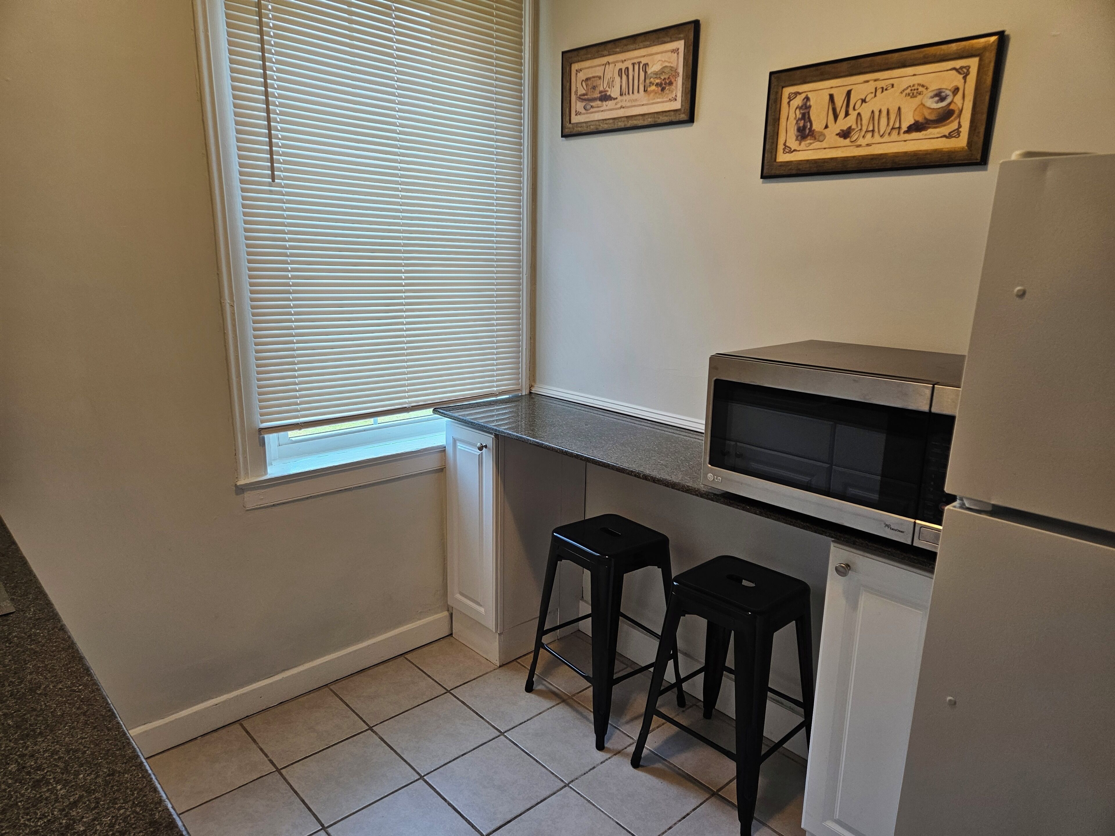 A kitchen with a microwave, two black stools, a refrigerator, and a window with blinds.