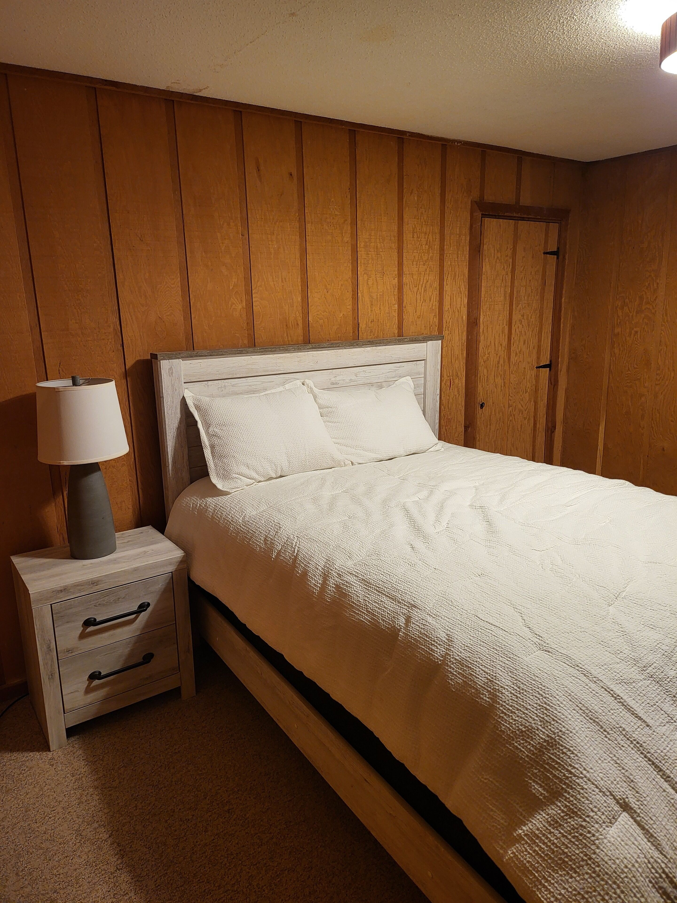 A bedroom with a wooden paneled wall, a single bed with white bedding, a nightstand with a lamp, and a door.
