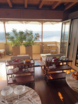 A balcony with a dining setup, a sofa, a table with a tray of food, and a view of the sea. - Entre Ciel et Mer Hotel (Bonifacio)