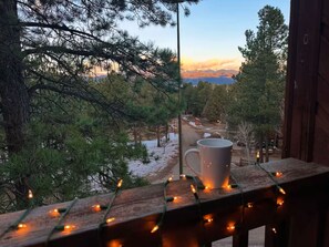 A wooden deck with a white mug and string lights, overlooking a snowy landscape and a cabin in the distance. - The Lifts Loft - walk to Ski/Bike lift (Angel Fire)