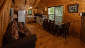 Ein Blockhaus-Interieur mit Holzboden, einer Couch mit gemusterten Kissen, einem Esstisch mit Stühlen, einer Küchenzeile mit Geräten und einem Fenster mit Jalousien.