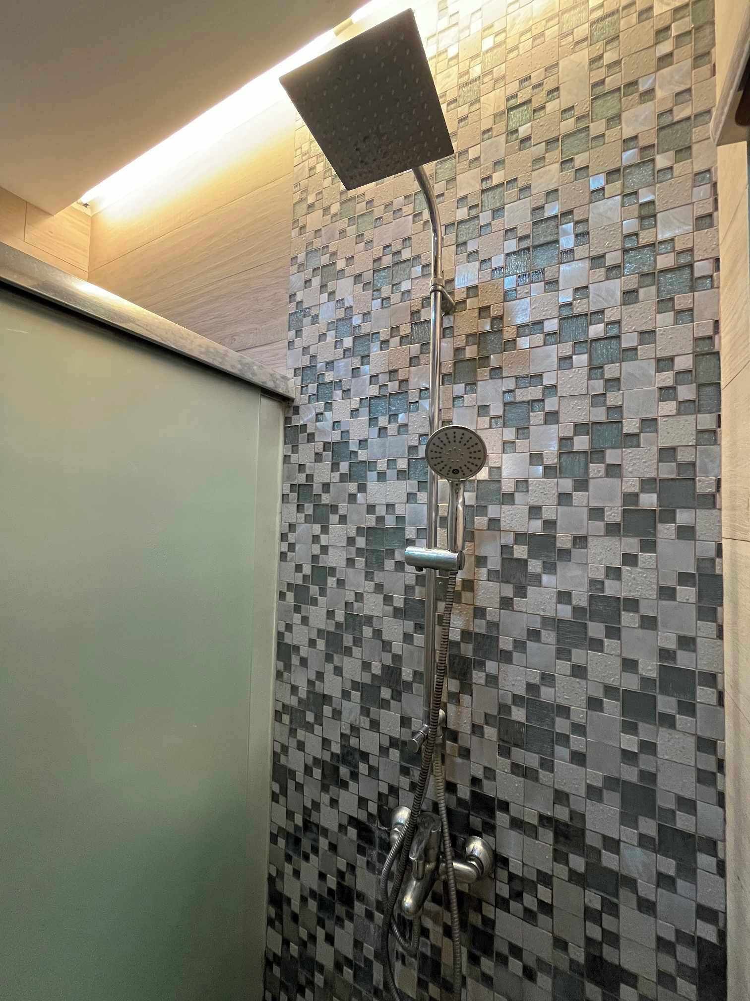 Une douche dotée d’un pommeau de douche carré et d’un pommeau de douche à main, le tout fixé sur un mur carrelé orné d’un motif en mosaïque.