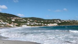 Ein Strand mit Wellen, einem großen Felsen und Häusern im Hintergrund.
